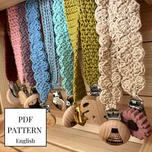 May include: A set of colorful crocheted straps with wooden and silicone clips. The straps are made in various colors and patterns, including pink, blue, green, and beige. The clips are attached to the straps and feature different designs, such as a flower, a heart, and a leaf. The text "PDF PATTERN English" is visible in the image.