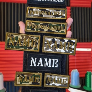 May include: A set of seven personalised name patches in various styles. The patches are black, white and camouflage with gold lettering. The patches are arranged in a vertical stack on a black background.