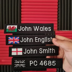 May include: Four black embroidered name tags with white text. The top tag has a Welsh flag and says "John Wales". The second tag has a Union Jack flag and says "John English". The third tag has a St. George's Cross flag and says "John Smith". The bottom tag has a Union Jack flag and says "PC 4685".