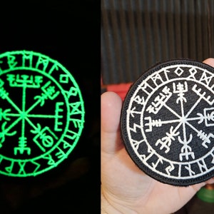 May include: A black and white embroidered patch featuring a compass design with runes around the edge. The compass has a central point with four arms extending out to the edges of the circle. The runes are in a circular pattern around the compass. The patch is on a black background.