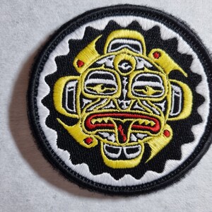 May include: A round black and white embroidered patch with a yellow and red stylized face in the centre.