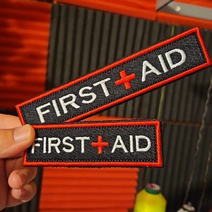 May include: Two black rectangular patches with the words "FIRST AID" in white, and a red cross symbol. The patches have a red border. One patch is slightly smaller than the other.