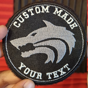May include: Black and white custom-made embroidered patch with a wolf head design. The text "CUSTOM MADE" is above the wolf and the text "YOUR TEXT" is below the wolf.