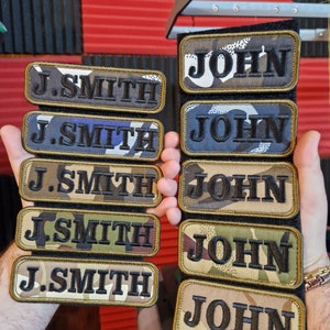 May include: Five camouflage fabric patches with the name "J. Smith" embroidered in black thread and five camouflage fabric patches with the name "John" embroidered in black thread.