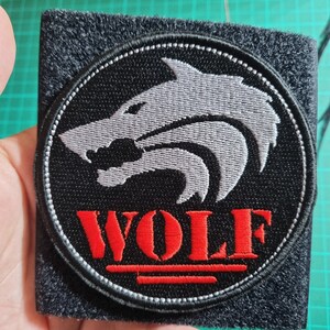 May include: A black and grey embroidered patch featuring a white wolf design and the word "WOLF" in red lettering.