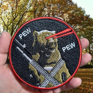 May include: A black and green embroidered patch featuring a cartoon character holding two rifles. The character has a red laser sight on the rifle. The patch reads "PEW" on either side of the character.