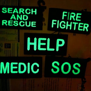 May include: Five black rectangular patches with glowing green text. The patches read: "SEARCH AND RESCUE", "FIRE FIGHTER", "HELP", "MEDIC", and "SOS". These patches are likely for emergency services personnel.