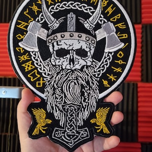 May include: Black and white embroidered patch featuring a skull wearing a horned helmet and holding axes. The skull has a long beard and is encircled by a circle of yellow runes. Below the skull is a silver hammer with a knotwork design and two yellow birds on either side.
