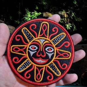 May include: A round embroidered patch with a black background and a red, yellow, and white sun face design. The sun face has a smiling mouth, two eyes, and a nose.