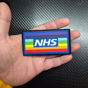 May include: A rectangular patch featuring a horizontal rainbow stripe pattern with a central blue rectangle containing the white text NHS. The patch has a black hook-and-loop backing border and is constructed from a smooth, flexible material.