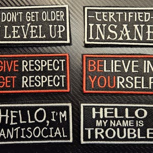 May include: Six black and white embroidered patches with various sayings. The patches include "I don't get older I level up", "Certified Insane", "Give Respect Get Respect", "Believe in Yourself", "Hello, I'm Antisocial", and "Hello My Name is Trouble".