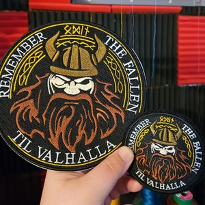 May include: Two embroidered patches featuring a Viking warrior design. The larger patch is black with gold trim and reads "Remember the Fallen Til Valhalla". The smaller patch is black with gold trim and reads "Remember the Fallen Til Valhalla".