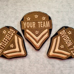 May include: Three embroidered patches in the shape of a shield with a chevron design. The patches are green with white stitching and feature the text "Battlefield", "Your Team", and "Division 722".