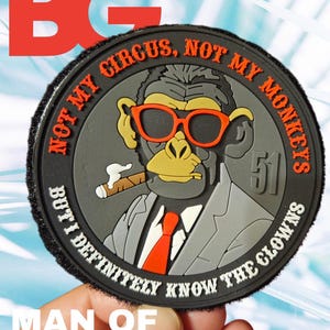 May include: A circular patch with a cartoon monkey wearing orange sunglasses, a suit, and a red tie, holding a cigar. The text around the edge reads "NOT MY CIRCUS, NOT MY MONKEYS BUT I DEFINITELY KNOW THE CLOWNS." The text "MAN OF THE YEAR" is at the bottom.