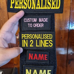 May include: Black velcro patch with five rows of white text. The top row reads "PERSONALISED". The second row reads "CUSTOM MADE TO ORDER". The third row reads "PERSONALISED IN 2 LINES". The fourth row reads "NAME" in red. The fifth row reads "NAME" in yellow.