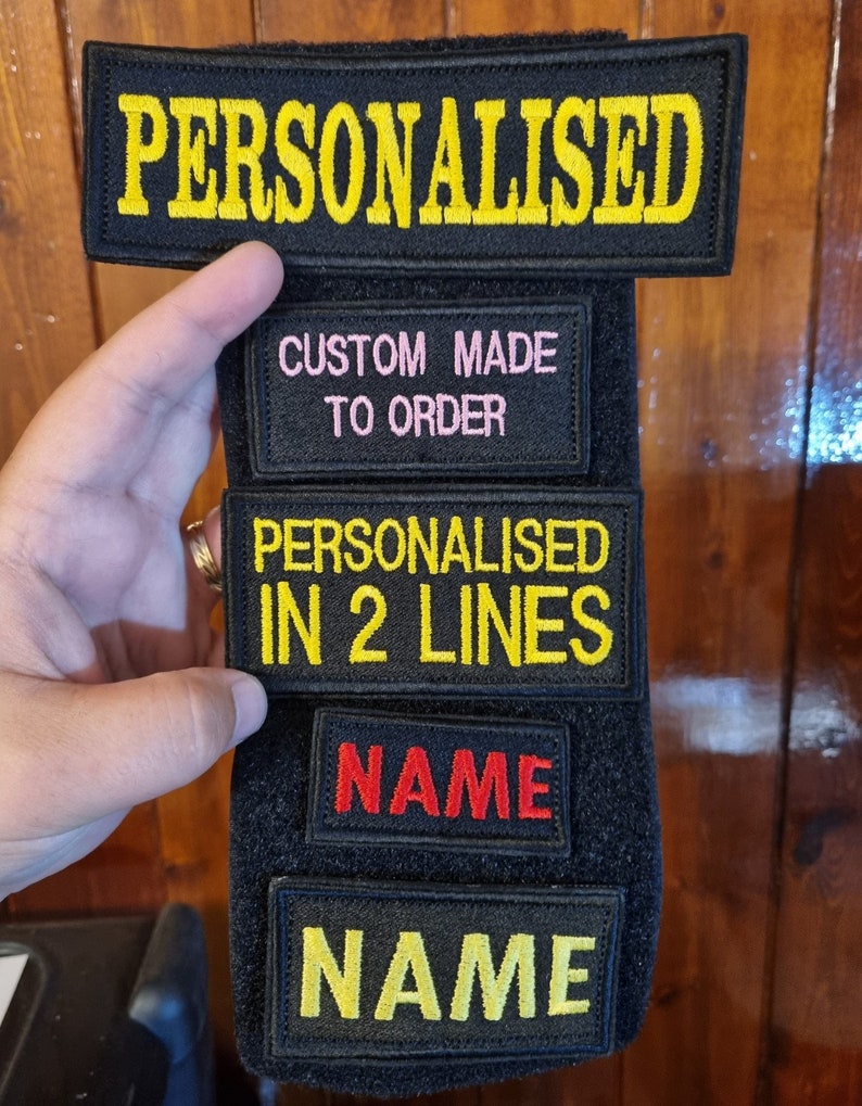 May include: Black velcro patch with five rows of white text. The top row reads "PERSONALISED". The second row reads "CUSTOM MADE TO ORDER". The third row reads "PERSONALISED IN 2 LINES". The fourth row reads "NAME" in red. The fifth row reads "NAME" in yellow.