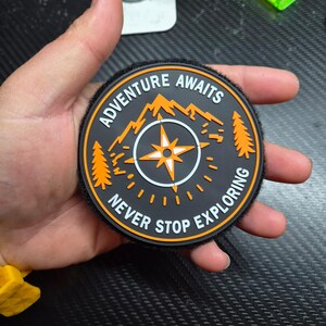 May include: A circular black patch featuring an orange compass rose in the centre, flanked by two orange pine trees and a mountain silhouette. The text ADVENTURE AWAITS is printed at the top, and NEVER STOP EXPLORING is printed at the bottom.