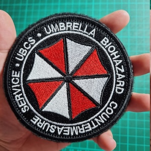 May include: A black and white embroidered patch with a red umbrella and the text "UBCS UMBRELLA BIOHAZARD COUNTERMEASURE SERVICE".