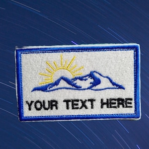 May include: White embroidered patch with a blue border. The patch features a yellow sun rising over blue mountains. The text "YOUR TEXT HERE" is embroidered in black below the mountains.