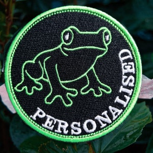 May include: A black and green embroidered patch with a frog design and the text "PERSONALISED" around the edge.