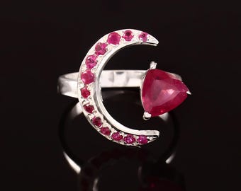 Könnte beinhalten: Ein silberner Ring mit einem Halbmond-Design, besetzt mit kleinen roten Edelsteinen. Ein größerer roter Edelstein ist an der Seite des Rings angebracht.