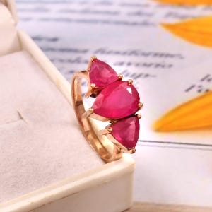 May include: A rose gold ring featuring three pear-shaped ruby gemstones. The ring is set on a delicate band with the stones arranged in a staggered pattern.