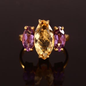 Könnte beinhalten: Ein goldener Ring mit drei Edelsteinen. Der Mittelstein ist ein großer, marquiseförmiger, goldgelber Edelstein, flankiert von zwei kleineren, oval geschliffenen, lila Edelsteinen. Der Ring hat ein poliertes Goldband und einen dunklen Hintergrund.