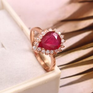 Può includere: Un anello in oro rosa con una pietra preziosa di rubino a forma di pera circondata da un alone di diamanti più piccoli.