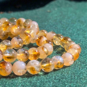 Könnte beinhalten: Nahaufnahme eines Armbands aus durchscheinenden, runden Perlen. Die Perlen zeigen eine Mischung aus hellgelben und bernsteinfarbenen Farbtönen mit unterschiedlichen Transparenzgraden. Das Armband liegt auf einer grünen Oberfläche, was die warmen Farbtöne der Perlen hervorhebt.