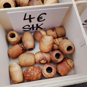 Op de afbeelding: Houten kralen in diverse vormen en maten, tentoongesteld in een houten kist. De kralen zijn lichtbruin met natuurlijke houtnerfpatronen. Sommige kralen hebben gaten. Het bord in de kist leest "4€".