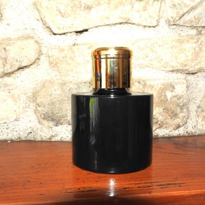May include: A black, cylindrical bottle with a shiny gold cap. The bottle is on a wooden surface, with a stone wall behind it. The bottle is likely for perfume or cologne.