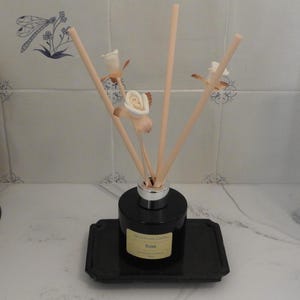 May include: A black glass bottle with a silver lid and wooden sticks, filled with a rose scented diffuser oil. The bottle is sitting on a black tray and has a label that reads "Rose" and "Luxury Wood Diffuser Oil".