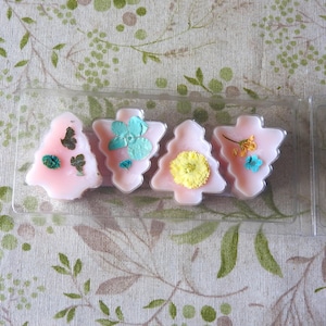 May include: Four pink, tree-shaped wax melts with embedded dried flowers in a clear plastic container. The flowers are various colours, including green, blue, yellow, and orange. The background is a floral patterned fabric.