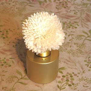 May include: A gold-coloured cylindrical vase with a cream-coloured, flower-shaped diffuser. The diffuser has many petals. The vase is on a floral patterned surface. The vase is approximately 8 cm tall.