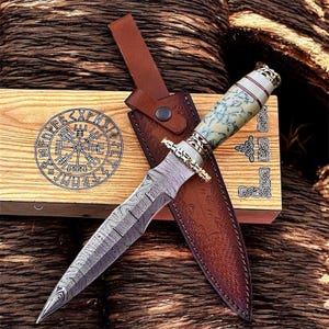 Op de afbeelding: Een Damascus stalen dolk met een patroon op het lemmet en een decoratief handvat met blauw en wit marmer. De dolk zit in een bruine leren schede, met een houten kist op de achtergrond. De kist heeft een cirkelvormig ontwerp.