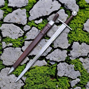 Puede incluir: Una espada de acero con hoja recta de doble filo, guarda plateada y empuñadura marrón oscuro rematada con un pomo esférico. Junto a la espada hay una vaina marrón oscuro a juego con una contera y una boquilla de metal plateado.