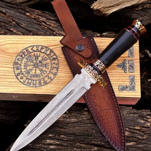 May include: A decorative dagger with a patterned blade and a black handle with gold accents. The dagger is in a brown leather sheath, resting on a wooden box with a compass rose design. The box and sheath are on a wooden surface.