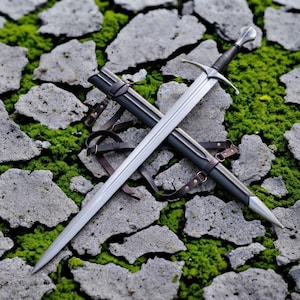 May include: A silver-toned metal sword with a dark handle and a cross-shaped guard, positioned diagonally across a matching dark scabbard. The scabbard features brown leather straps and buckles, with a pointed silver tip at the bottom end.