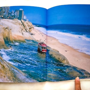 May include: A vibrant photograph of a magazine spread featuring a red cable car emerging from the ocean onto a sandy beach. The scene includes buildings, cliffs, and a bright blue sky, creating a surreal landscape.