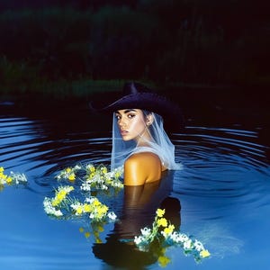May include: A person wearing a black cowboy hat and a white veil, standing in water. Yellow and white flowers float on the water's surface. The background is a dark, blurred landscape.