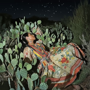 May include: A person wearing a colorful patterned dress with floral and geometric designs, lying among green cactus plants under a starry night sky. The dress features red, yellow, and orange accents.