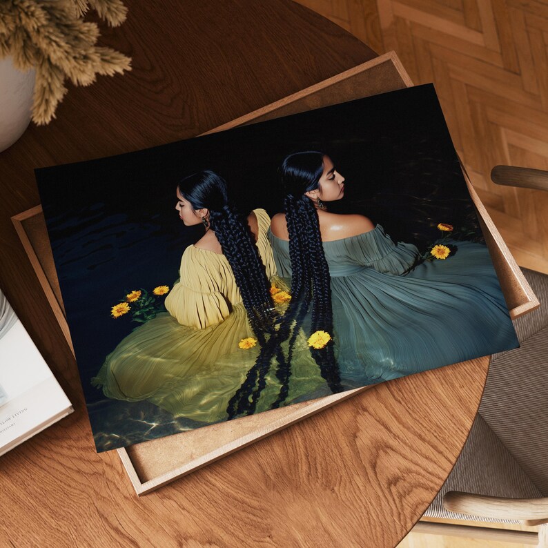 May include: A framed art print featuring two women with long, braided black hair, wearing off-the-shoulder gowns in yellow and teal. Yellow flowers float in the water around them. The print is displayed on a wooden surface.