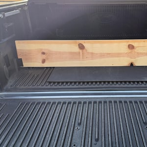 May include: A wooden plank in the bed of a pickup truck. The plank is light brown and has a natural wood grain. The truck bed is lined with a black rubber mat.