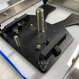 May include: A black metal plate with a silver metal rod in the center. The plate has a black knob on the right side and a black strap on the left side. The plate is attached to a silver metal frame.