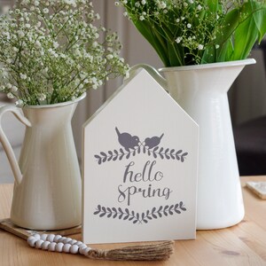 Könnte beinhalten: Ein weißes Holzschild mit dem Text "hello spring" in grauer Schrift. Zwei graue Vögel sitzen auf einem Ast über dem Text. Das Schild ist mit grauen Blättern verziert. Das Schild steht auf einem Holztisch mit zwei weißen Krügen, die mit weißen Blumen gefüllt sind.