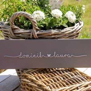 Könnte beinhalten: Ein graues Holzschild mit dem Text "dominik ♥ laura" in weißer, kursiv geschriebener Schrift.