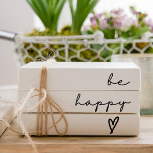 Könnte beinhalten: Ein Stapel aus drei weißen Büchern mit den Worten "be happy" und einem gezeichneten Herzen auf der Vorderseite. Die Bücher sind mit Schnur zusammengebunden.