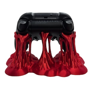 May include: A black gaming controller rests on a vibrant red stand. The stand has a unique, flowing design, with a glossy finish. The controller is a standard size, with buttons and joysticks visible. The overall aesthetic is modern and eye-catching.