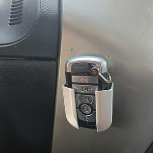 May include: A silver car key with a white key fob holder. The key has several buttons, including a lock, unlock, and panic button. The key is attached to a silver ring.