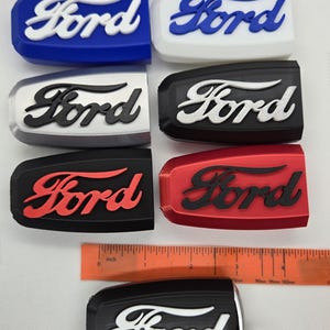 May include: Six different colored key fobs with the Ford logo in white, black, and red. The key fobs are made of plastic and have a smooth finish. The key fobs are arranged in two rows of three. A black key fob with a white Ford logo is shown on a ruler with inch markings.